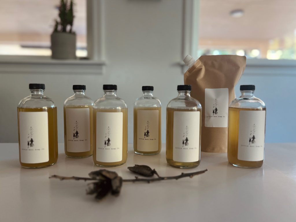 Little Root Soap Co liquid soaps in glass bottles and a kraft refill pouch on a kitchen counter